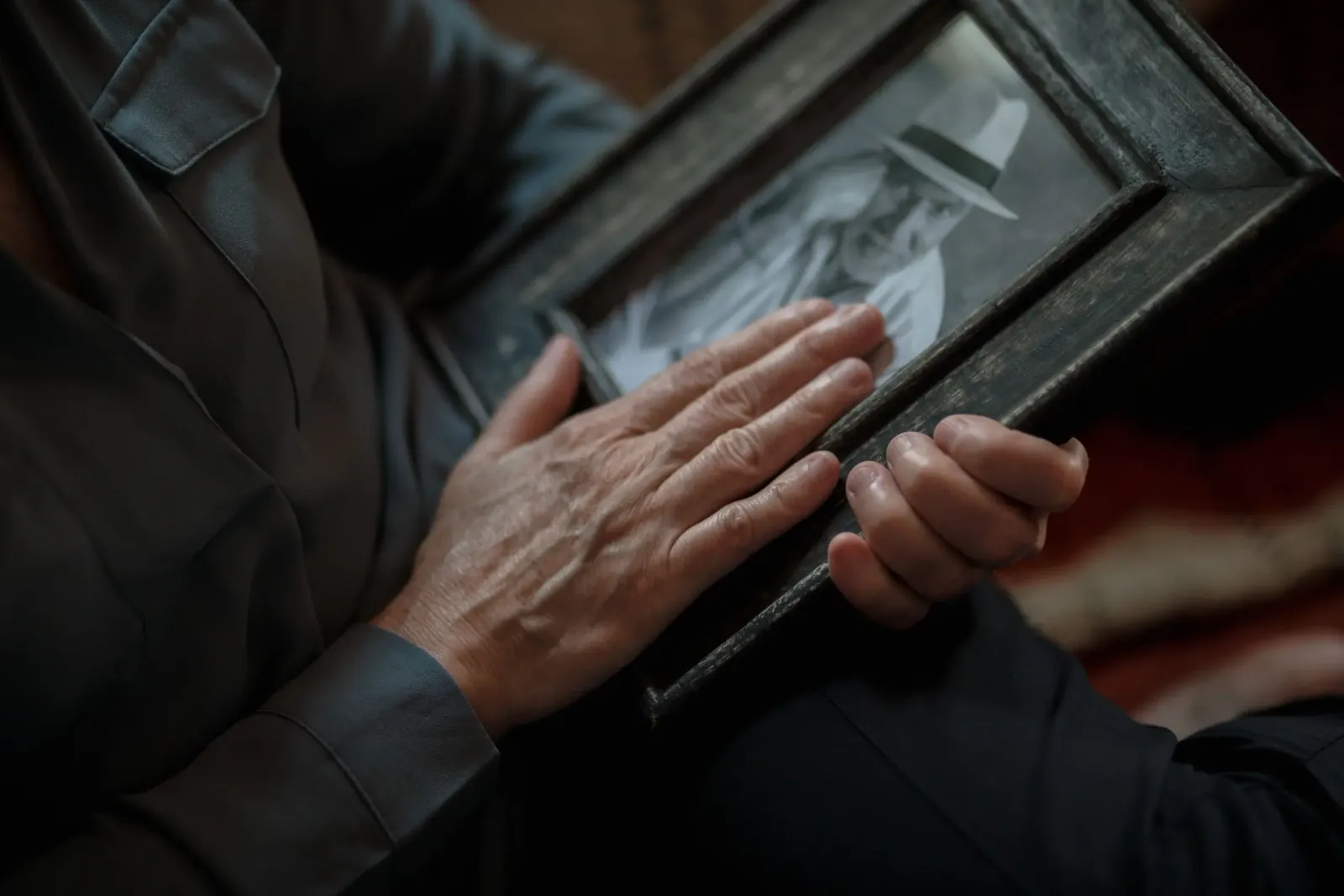 An old, weathered hand rests on a framed black and white portrait of a man in a hat