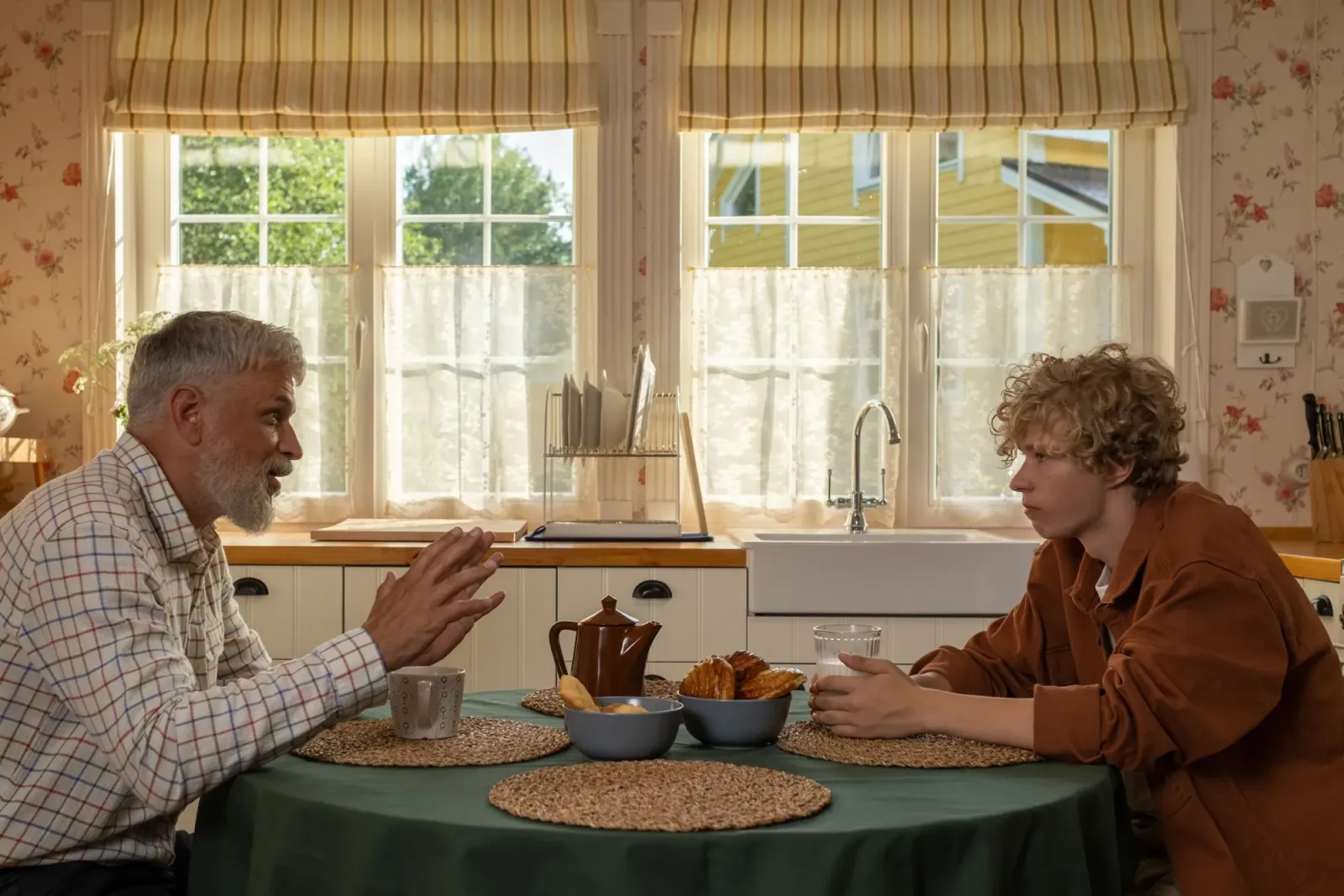 A grandfather with a grey beard leans across a kitchen table in warm window light, telling a story to his teenage grandson, with a teapot and pastries between them